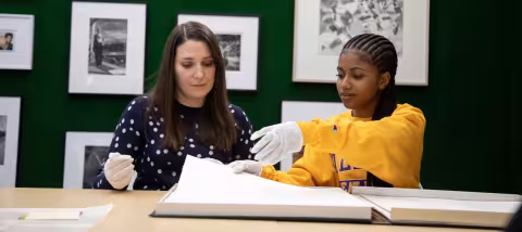 A student intern and a professional archivist, both wearing white cotton gloves, sit at a table and examine a piece of artwork together.
