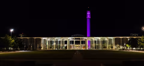 The UAlbany carillon tower lit up with purple lights and the Academic Podium buildings lit up with white lights at night.