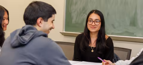 A student seated in front of a chalkboard smiles at another student during a class discussion.