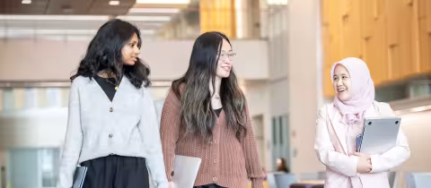 Three graduate students smile and chat as they walk through UAlbany's ETEC building carrying their laptops.