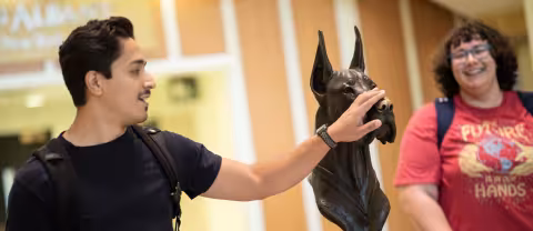 A student reaches out to touch the nose of a bronze Damien the Great Dane statue while another student looks on and smiles.