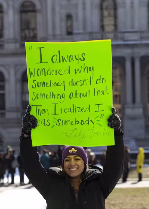 Student holds placard with quote from Lily Tomlin that reads "I always wondered why somebody doesn't do something about that. Then I realized I was somebody."
