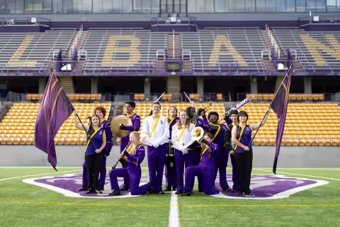 An ensemble of marching band members, two colorguard with flags, two drum majors, two percussionists, a clarinetist, and two trumpet players pose for a group photo in the UAlbany Football stadium. They are all wearing the UAlbany marching band uniform, which is mostly purple with gold accents. The drum majors' uniforms have purple pants with a white coat and gold accents.