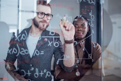 Two students writing mathematical formulas on a glass board