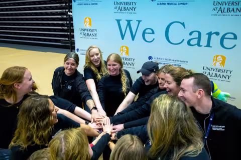 A group of people forming a hand stack in front of a We Care sign in the UAlbany gym.