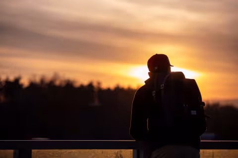 A UAlbany student with a backpack stands gazing at a vibrant sunset over the horizon.
