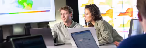Two researchers work on laptops inside a lab space where maps are projected on screens around them.