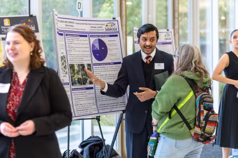 A student presenter gestures toward his research poster while speaking with a Showcase attendee.