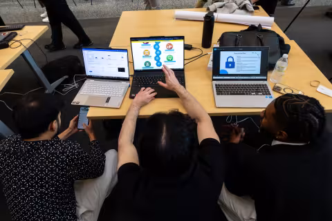 Three students sit at a table and collaborate on a presentation using their laptops.