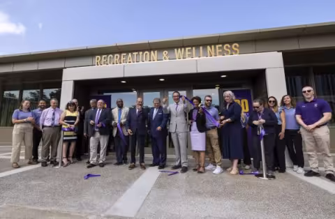 A line of smiling people cut a purple ribbon in front of a building with the words "Recreation & Wellness" above the doors.