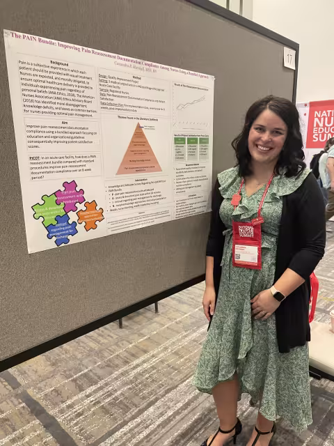 Marshall, wearing a green sundress and black cardigan, stands beside her poster at the National Nurse Education Summit. Her poster is titled “The PAIN Bundle: Improving Pain reassessment Documentation Compliance Among Nurses Using a Bundled Approach.” The poster is tacked to a gray display board. 