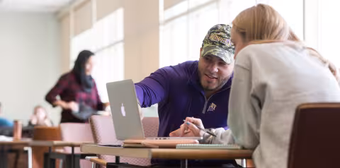 Two students sit at a table inside a study space. One student gestures toward a laptop screen while the other takes notes.