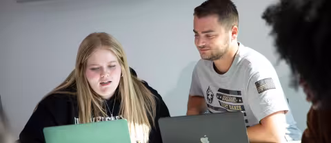 Two students sit together at a table with laptops open in front of them. One student smiles and looks on as the other student gestures and talks.