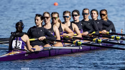 A team of 8 rowing women are on a rowing boat wearing purple uniforms with logos of the University at Albany on them