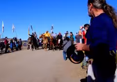 A group of people stand in a circle, some holding poles and some playing small hand tambourines.