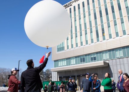 A man releases a large white weather balloon in front of about a dozen people standing outside ETEC at UAlbany.