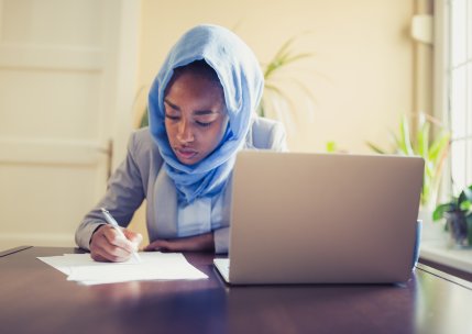 A woman sits at a table with a laptop in front of her. She uses paper to take notes.