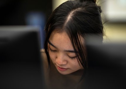 A student sits behind a desktop computer screen, looking down while working.