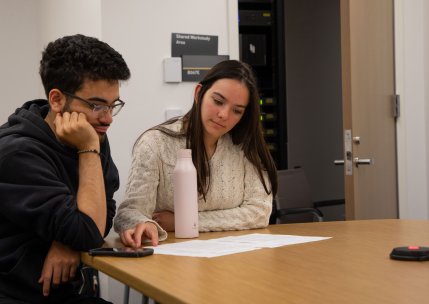 Two students sit at a table inside a tutoring space, looking at paper arrayed before them.