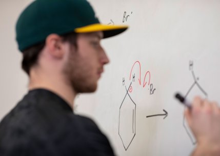 A student wearing a baseball hat works on a chemistry problem on a whiteboard.
