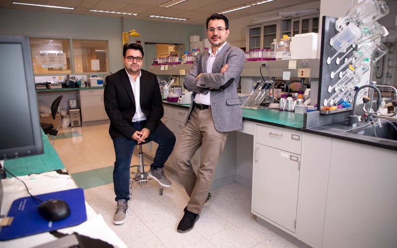 Two men pose for a portrait in a chemistry lab. One is seated, wearing a black blazer and jeans; the other is leaning against a lab bench wearing a gray blazer and khaki pants. Various glass and plastic containers are on the bench and shelf behind them.