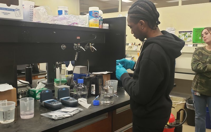 A young man wearing a black hoodie and blue latex gloves stands at a lab bench covered in various instruments used to detect legionella presence. The lab bench is black and the walls in the background are white. A young woman in a camo print shirt looks on. 