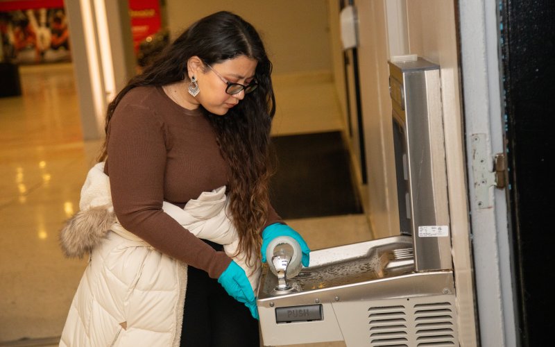 A young woman with long dark hair wearing glasses, a brown sweater, blue latex gloves and white winter coat tied around her waist fills a plastic water collection bottle at a silver water fountain. 