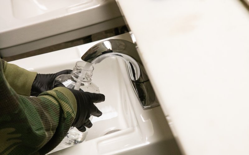 A person wearing a camo print long sleeved shirt and black latex gloves holds a clear plastic water bottle under a silver faucet to collect a water sample. The sink is white porcelain. 