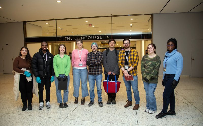 Nine smiling people, five women and four men, stand together for a group portrait under a sign that reads “The Concourse” at Empire State Plaze. Several are wearing latex gloves used to safely collect water samples. – I 