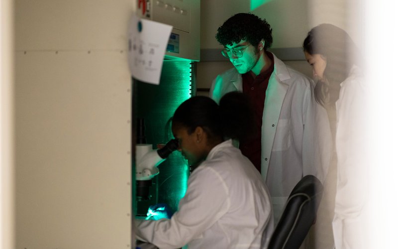Three students are pictured wearing white lab coats in a darkened room. One is seated, looking into a microscope which is emitting a green glow.
