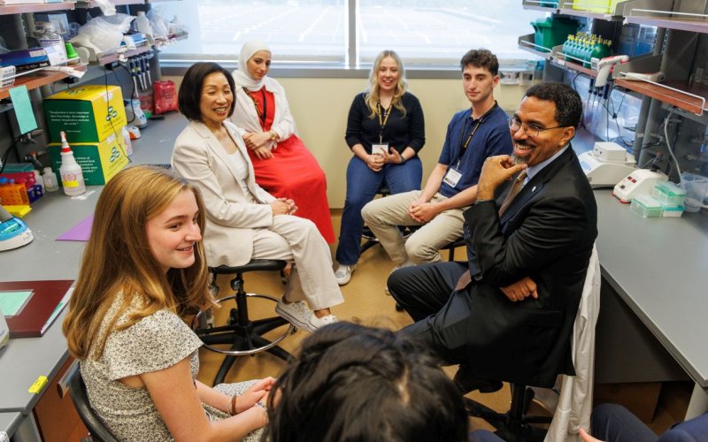 Seven smiling people are seated in a circle in a lab having a conversation.