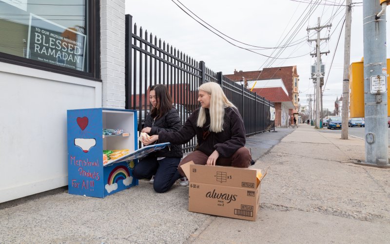 Two young women crouch on a sidewalk next to a blue metal box which they are stocking with a variety of period products.