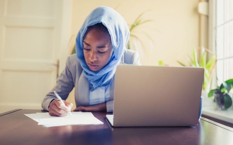A woman sits at a table with a laptop in front of her. She uses paper to take notes.