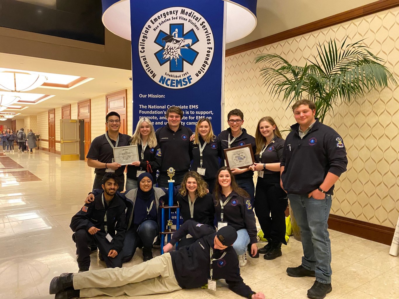 Twelve Five Quad volunteers pose for a photo with plaques and a trophy in front of a National Collegiate EMS Foundation sign