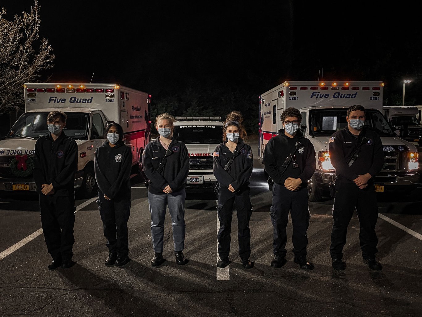 Six masked Five Quad volunteers pose for a photo in front of two ambulances and a paramedic's vehicle