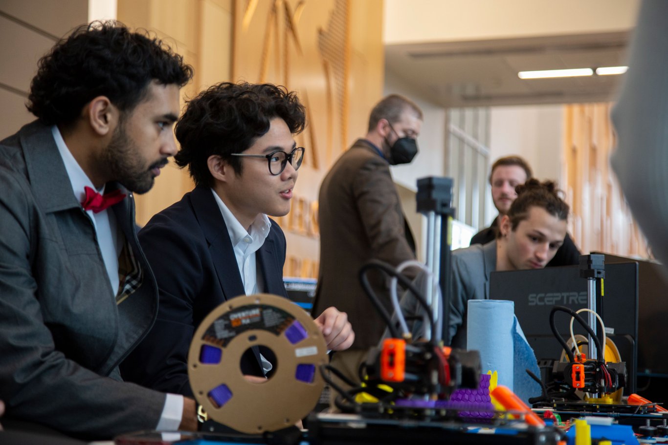 Two students in suits lean over a table with two 3D printers in front of them during UAlbany Showcase.