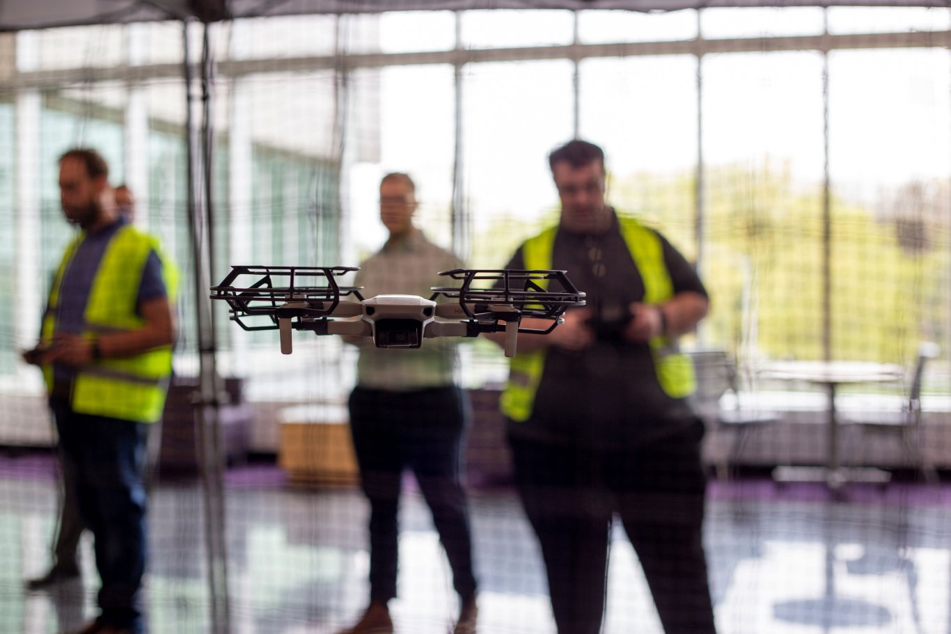 A quadcopter flies inside a netted arena, while a student in a yellow vest uses remote to control the drone at UAlbany Showcase.