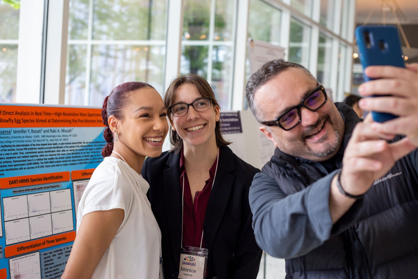 Two students pose for a photo as a professor takes a selfie with them in front of a poster at UAlbany Showcase.