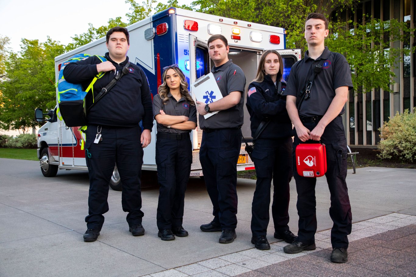 Five ambulance crew members in Five Quad uniforms and holding gear pose for a photo in front of an ambulance. 