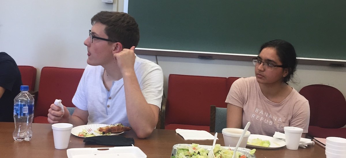 Two students enjoy some lunch as they listen to a peer's presentation.