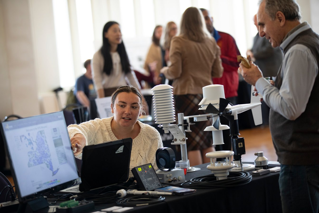 A woman sits at a table and points to a monitor displaying a data map of NYS as she shows a man her research at UAlbany's Research Resources Fair.
