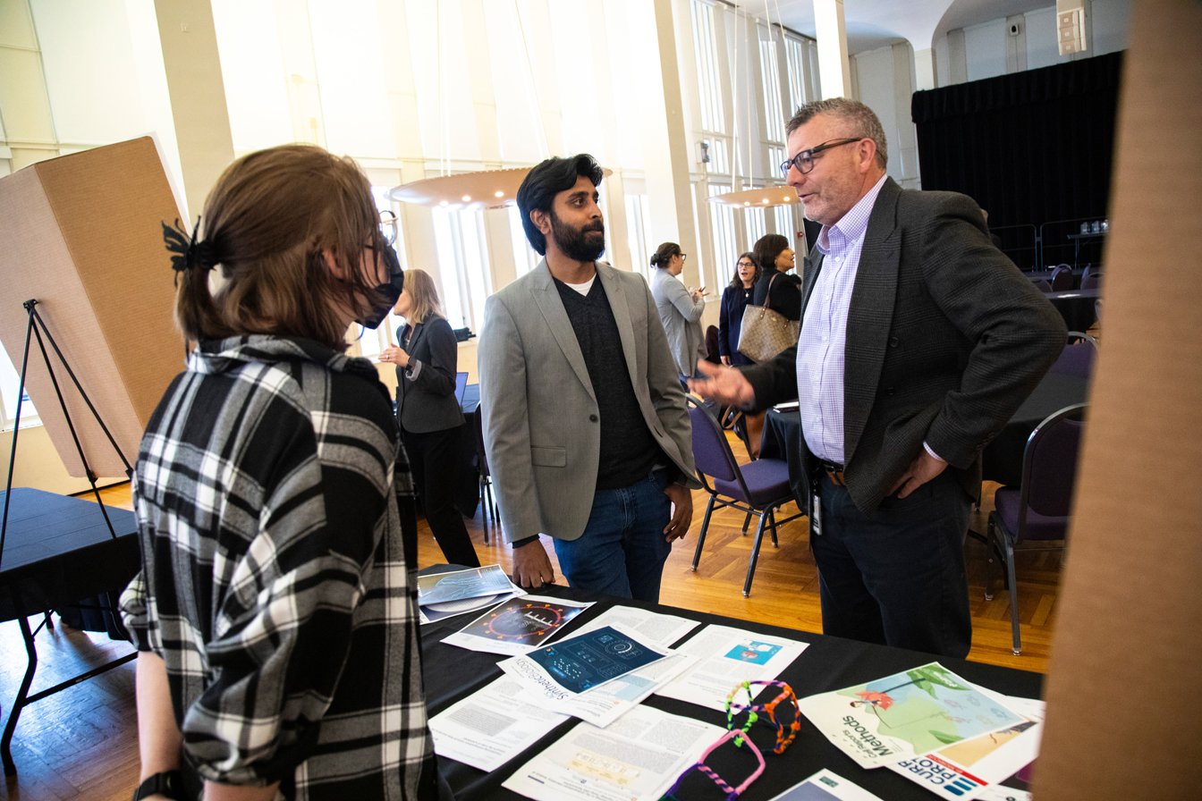 Three people stand around a table and discuss research in Albany's Campus Center Ballroom during the Research Resources Fair.  