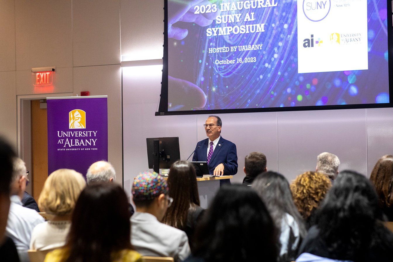 UAlbany President Havidán Rodríguez addresses a roomful of people at the SUNY AI Symposium. A screen behind him says "2023 Inaugural AI Symposium hosted by UAlbany, October 16, 2023" with the SUNY logo, the UAlbany logo and the AI plus logo.