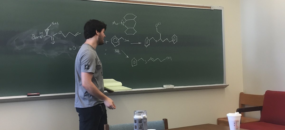 Team member Evan stands before a chalkboard featuring a sketch of a structural formula.