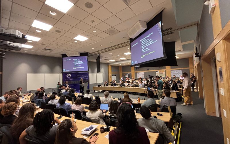 A presenter speaks at a podium inside a lecture hall full of conference participants. Two overhead screens display unreadable text.