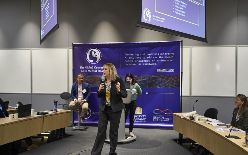 A woman gestures as she speaks into a microphone. She is standing in front of a GCAIMH Annual Summit banner.