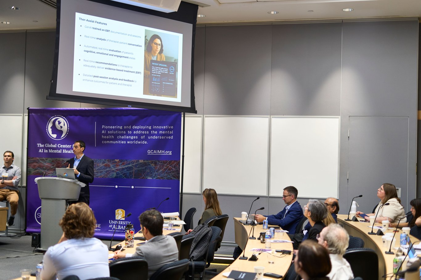 Audience members listen as a man presents a slideshow from a podium. He is standing in front of a GCAIMH Annual Summit banner.