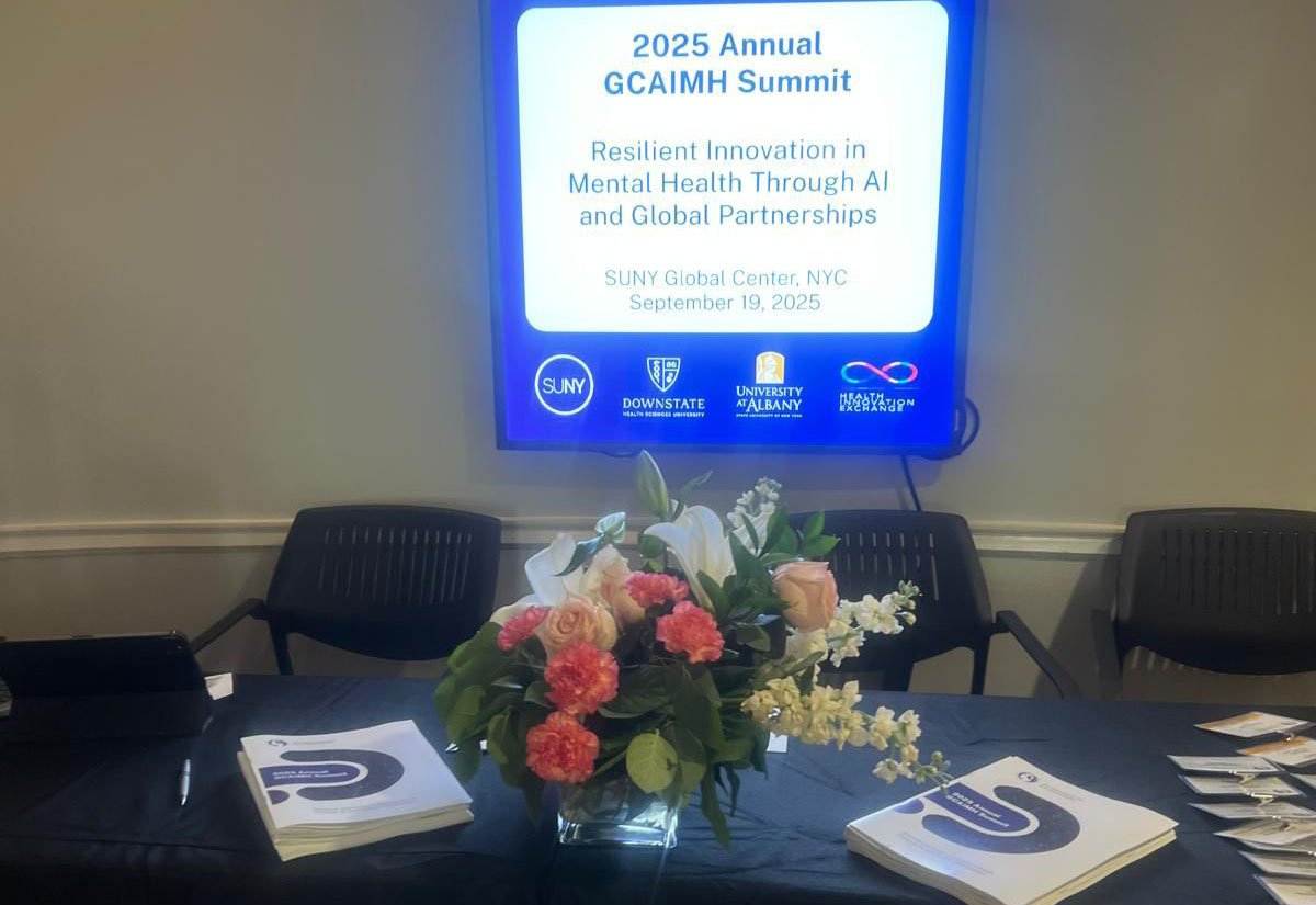 A registration table with pamphlets, flowers and a sign with the words, "2025 Annual GCAIMH Summit, Resilient Innovation in Mental Health through AI and Global Partnerships, SUNY Global Center, September 19, 2025," and the logos for SUNY, SUNY Downstate Health Sciences University, the University at Albany and the Health Innovation Exchange.
