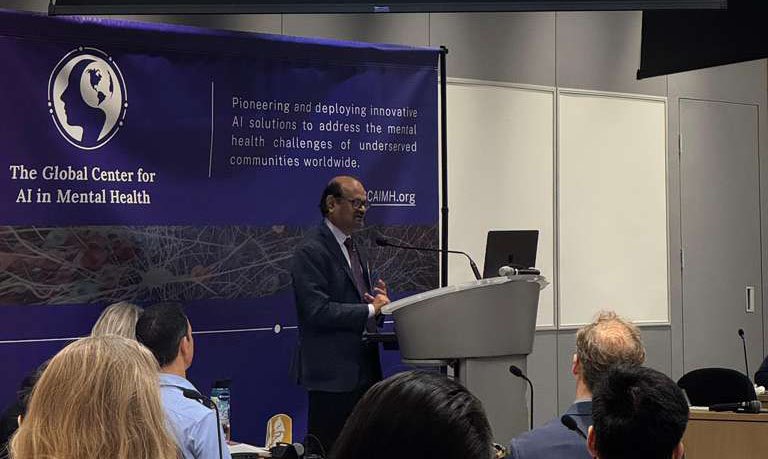A man speaks into a microphone mounted on a podium. He is standing in front of a GCAIMH Annual Summit banner.