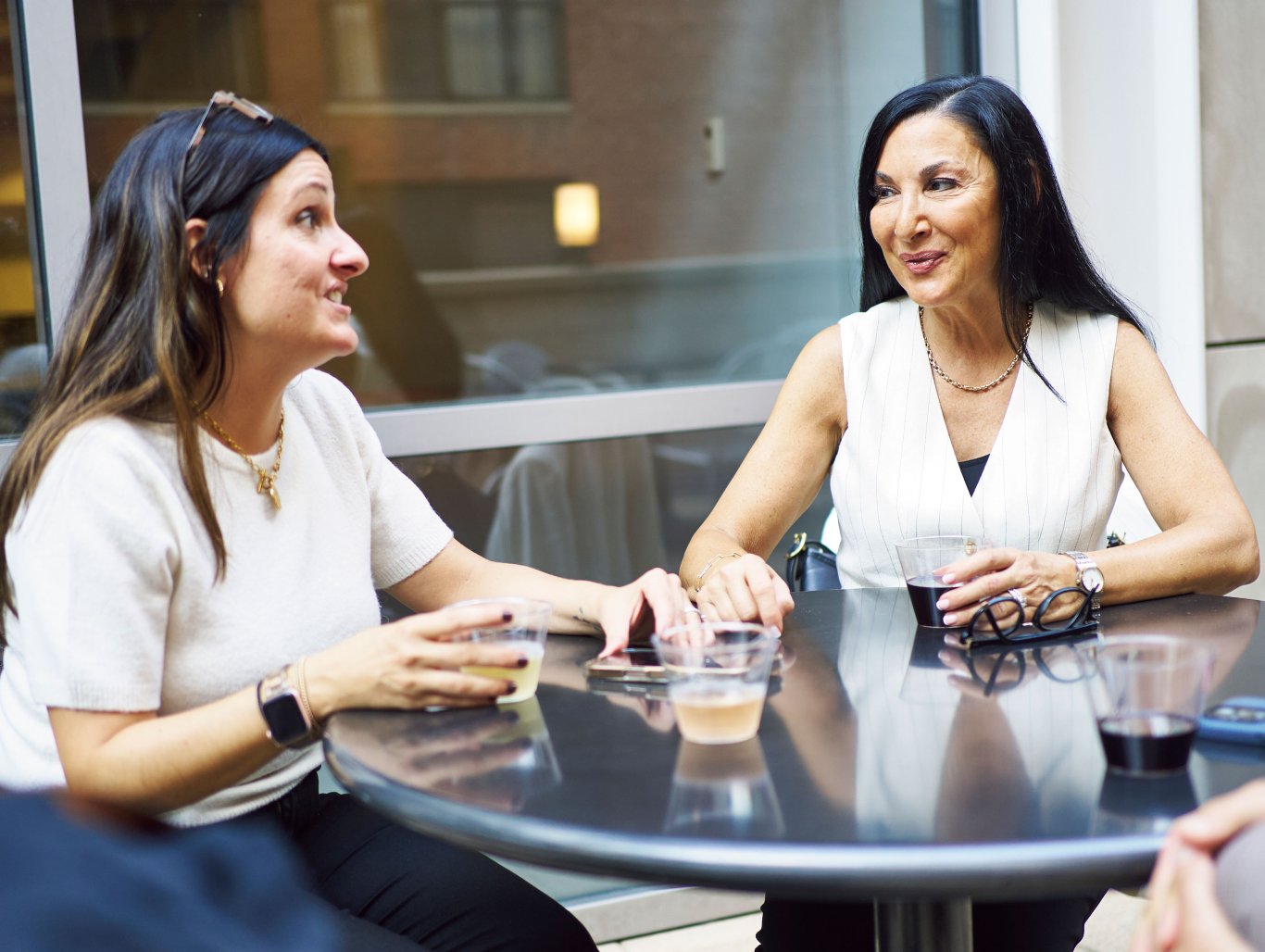 Two women sit at a table talking and smiling. On the table are drinks, a pair of eye glasses and a cell phone.